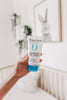 Hand holding a tube of Original Sprout Scrumptious Baby Cream in a blurred room setting